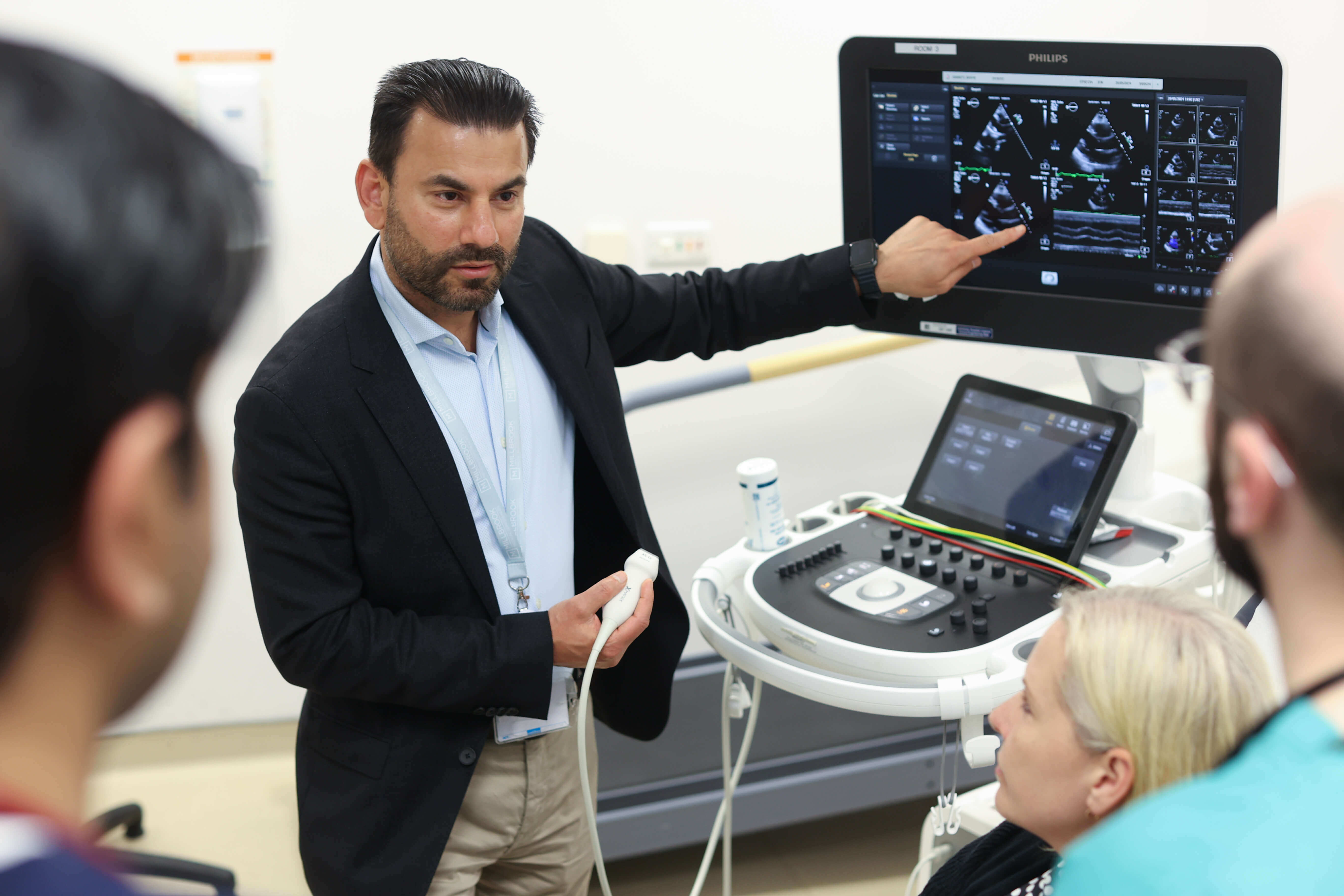 Man with beard points to scan image on ultrasound machine as he speaks to female patient and other doctors